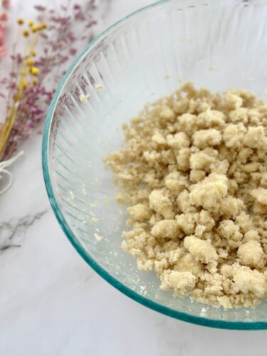 A glass bowl filled with a crumbly mixture, resembling a prepared dough or pastry base, on a marble surface with dried flowers in the background.