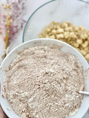A bowl of flour or dry ingredients in the foreground with a mixing bowl of dough in the background, decorated with dried flowers.