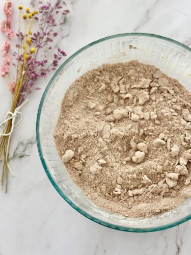 A glass bowl filled with beige flour or dry mix, surrounded by dried flowers on a marble surface.