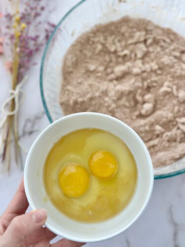 A person's hand holding a white bowl containing two cracked eggs, with a mixing bowl of dry ingredients in the background.