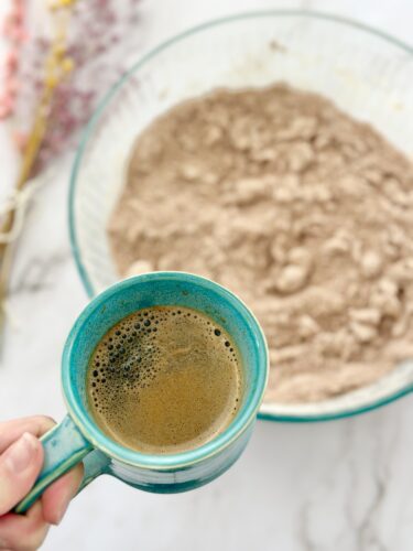 A hand holding a teal mug of coffee in the foreground, with a bowl of dry ingredients in the background.