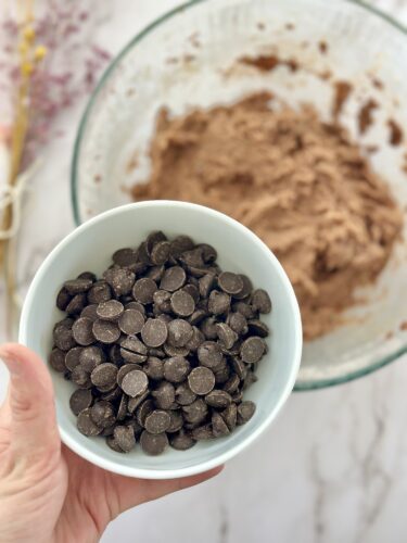 A hand holding a bowl of chocolate chips in front of a mixing bowl with brown dough in the background.