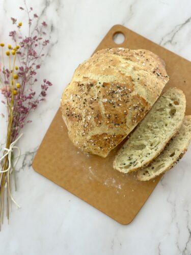 A freshly baked round loaf of bread, sliced to show the soft interior, resting on a wooden cutting board. Dried flowers are placed beside it.
