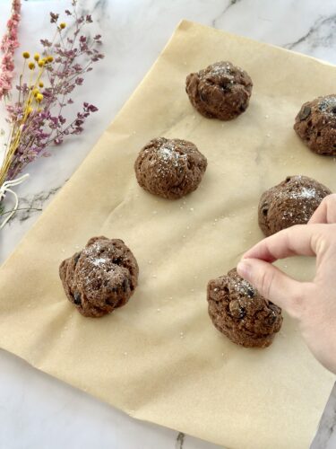 Six chocolate cookie dough balls on parchment paper with a hand sprinkling flour on top