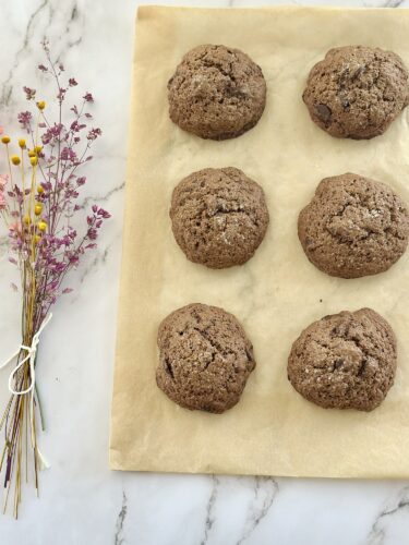 Freshly baked chocolate chip cookies arranged on parchment paper, with a bouquet of dried flowers on the side.