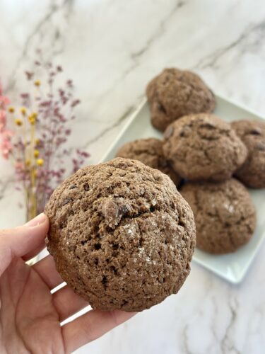 A hand holding a large chocolate cookie, with more cookies displayed on a plate in the background, surrounded by soft, decorative flowers.