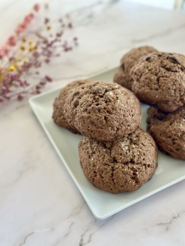 A plate of freshly baked chocolate cookies, placed on a marble countertop, with decorative dried flowers in the background.