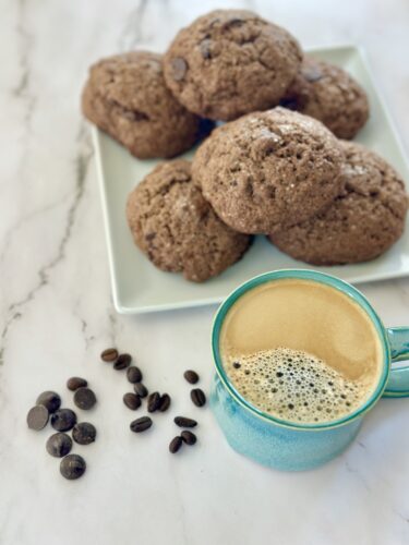 A plate of freshly baked chocolate chip cookies next to a blue coffee mug filled with frothy coffee, with scattered coffee beans on a marble surface.