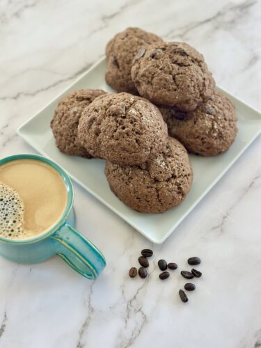 A plate of freshly baked chocolate cookies next to a cup of coffee and scattered coffee beans on a marble surface.