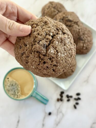A hand holding a large chocolate cookie with a plate of cookies and a cup of coffee in the background.