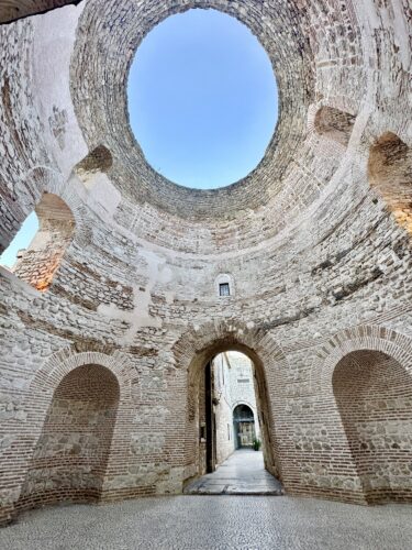 Interior view of a historic stone structure featuring a circular ceiling opening and arched doorways, with a view of a pathway beyond.