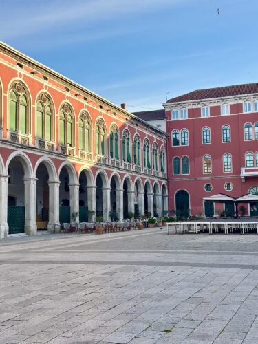 A scenic view of a historic plaza featuring red buildings with arches and green windows, set under a clear blue sky.