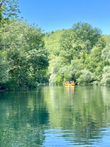A group of people paddling in a yellow raft on a calm river surrounded by lush green trees and a clear blue sky.