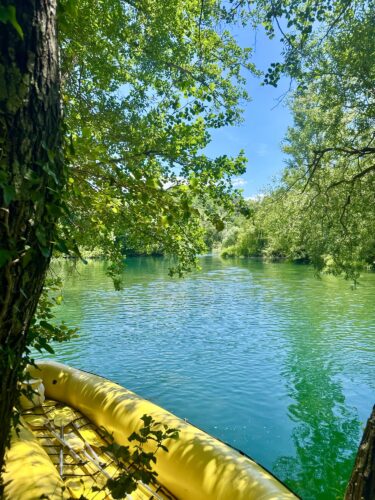 A peaceful view of a river surrounded by lush green trees, with a yellow inflatable boat partially visible in the foreground.