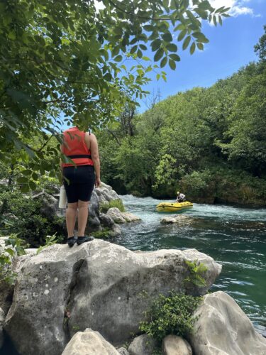 A person wearing a red life jacket stands on a rock by the river, looking at a yellow inflatable kayak floating downstream, surrounded by lush green trees and a clear blue sky.