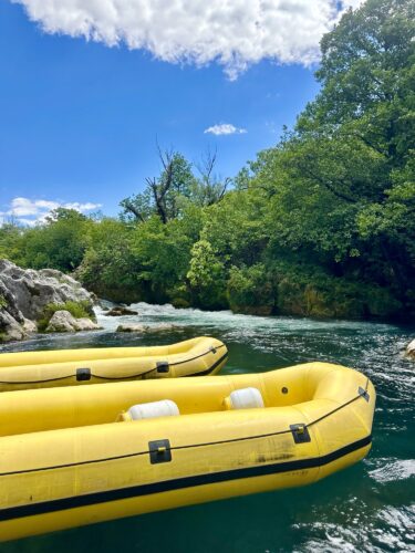 Two yellow inflatable rafts on a clear blue river surrounded by lush green trees and rocky shores under a partly cloudy sky.