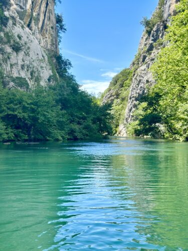 A serene view of a turquoise river flowing between steep, rocky cliffs and lush green trees under a clear blue sky.