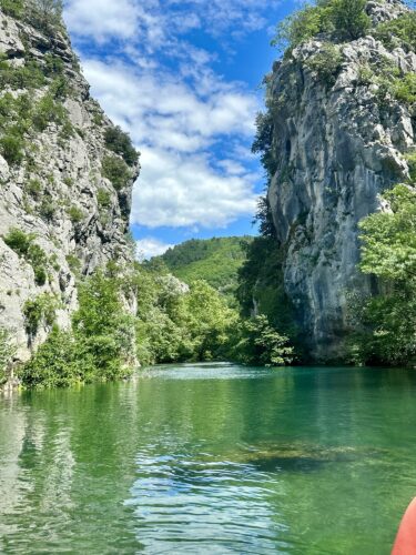 A serene river surrounded by tall rocky cliffs and lush greenery under a partly cloudy sky.