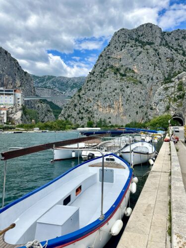 A scenic view of a marina with several boats docked on calm waters, surrounded by towering rocky cliffs and a cloudy sky.