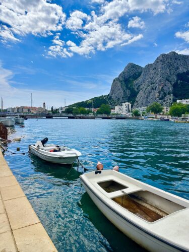 Two boats moored in a calm harbor with rocky mountains in the background under a partly cloudy blue sky.