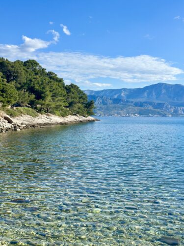 A serene coastal view featuring clear blue water, rocky shorelines, and lush greenery against a backdrop of mountains under a bright blue sky.