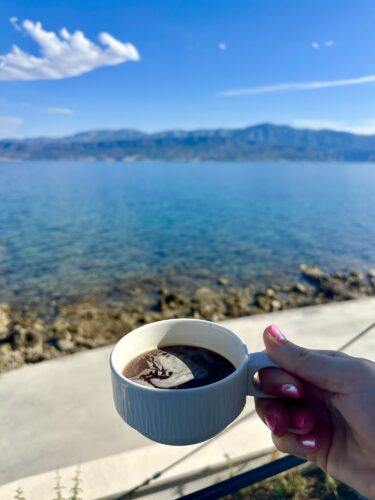 A hand holding a white cup of coffee, with a scenic view of a blue lake and mountains in the background under a clear sky.