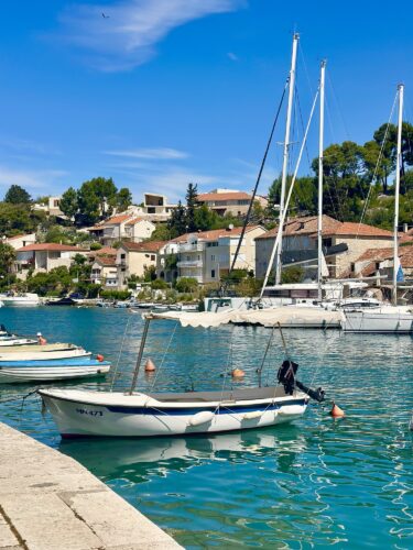 A small boat in a tranquil harbor with houses and trees in the background, under a blue sky.