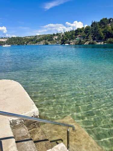 Clear turquoise water gently lapping against a stone pier, with a metal handrail leading down into the water, surrounded by lush greenery and distant hills under a blue sky.