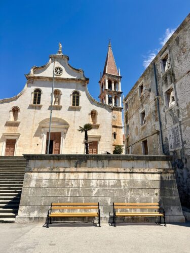 A historic church with a bell tower and a staircase leading up to its entrance, set against a clear blue sky. There are two wooden benches in the foreground and an adjacent weathered stone building.