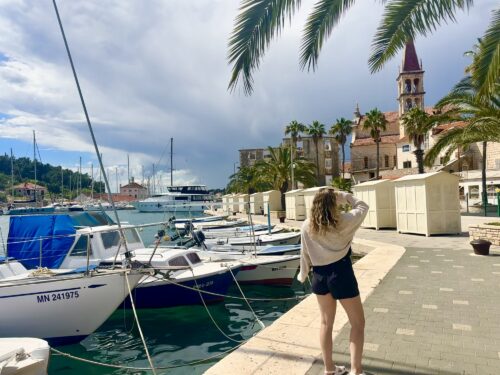 A woman standing near a marina, looking at the boats and waterfront, with palm trees and a church tower in the background under a partly cloudy sky.