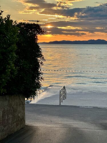Sunset over the water with orange and blue hues, viewed from a pathway lined with greenery and stairs leading down to the beach.