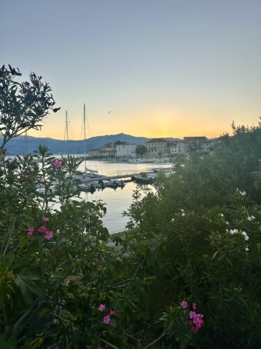 A serene waterfront scene at sunset featuring boats in a harbor, surrounded by lush greenery and blooming flowers with mountains in the background.