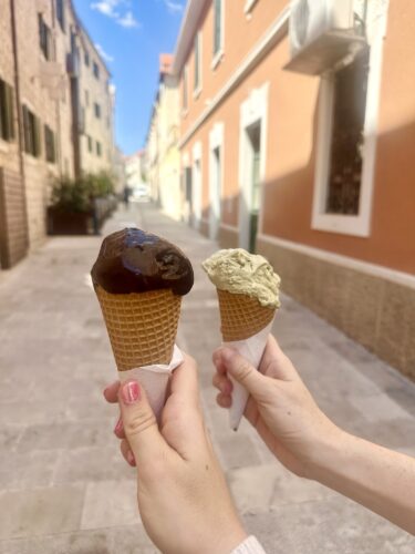 Two hands holding ice cream cones in a sunny street, one with chocolate ice cream and the other with a green flavor.