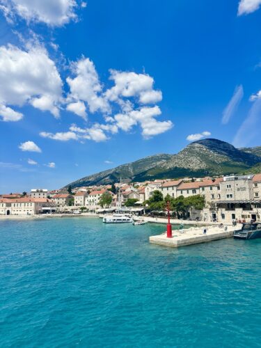 A scenic view of a coastal town with colorful buildings along the shore, featuring boats in the turquoise water and mountains in the background under a partly cloudy blue sky.