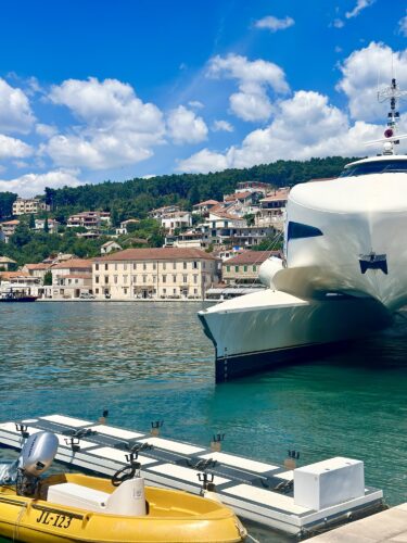 A luxury yacht docked in a marina with a scenic background of houses on a hillside under a partly cloudy sky.