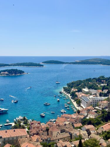 A panoramic view of a turquoise bay with several boats, surrounded by green islands and a coastal town with terracotta-roofed buildings under a clear blue sky.