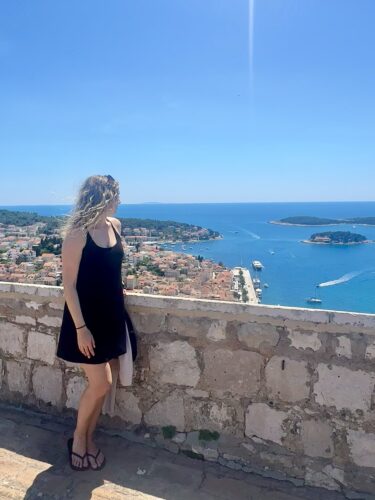 A woman in a black dress stands on a stone wall overlooking a coastal town and blue waters under a clear sky.