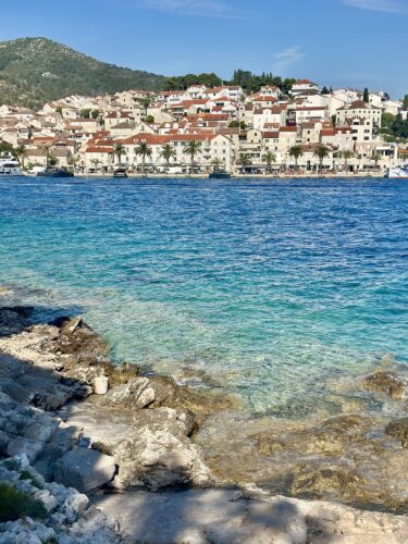 View of a coastal town with white buildings and red rooftops, surrounded by clear blue water and rocky shoreline.
