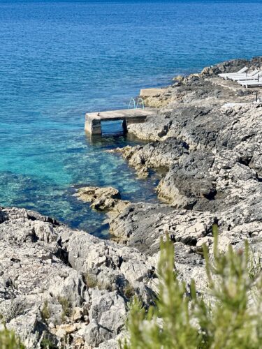 A scenic view of a rocky coastline with clear turquoise water, featuring a small concrete jetty and steps leading into the water.