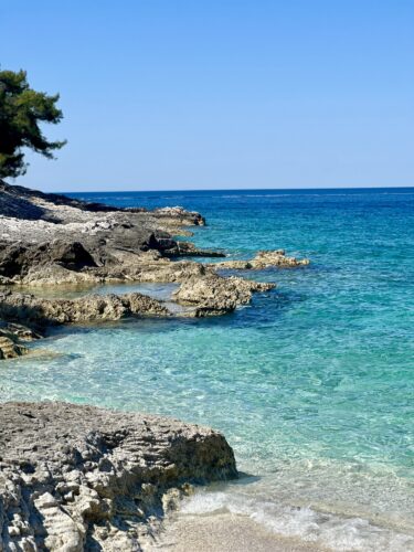 A clear blue sea gently lapping against rocky shorelines under a bright blue sky.