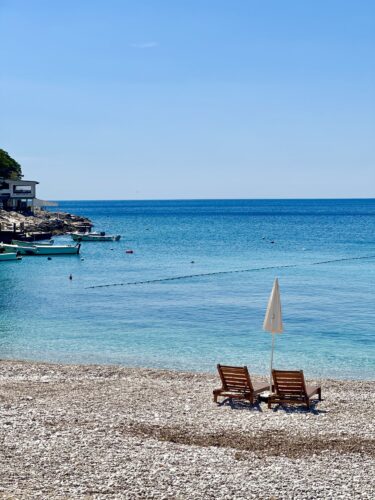 Two wooden lounge chairs under a white umbrella on a pebbled beach beside a calm blue sea.