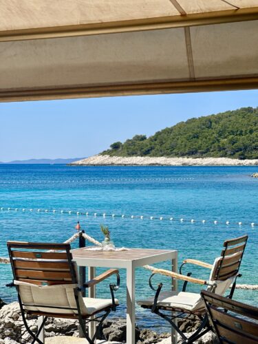 A serene coastal view from a shaded area with two chairs and a table overlooking clear blue waters and distant green hills.