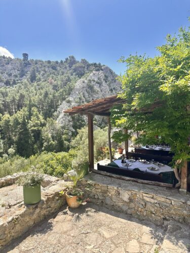 A scenic outdoor dining area with a wooden pergola, surrounded by lush greenery and mountains in the background. A table is set for a meal, adding to the charming atmosphere.