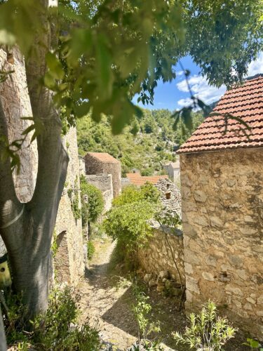 A narrow, winding path between rustic stone buildings, lined with greenery, leading into a mountainous area under a blue sky.