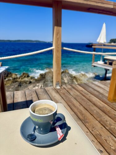 A cup of coffee on a table with a scenic view of the ocean and sailboat in the background, on a wooden deck.