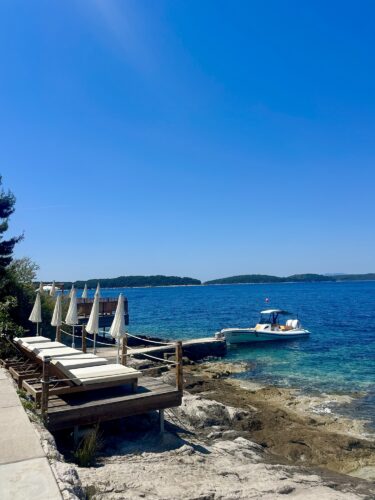 A scenic view of a calm blue sea with a small boat docked, surrounded by rocky shorelines and lounge chairs with umbrellas on a sunny day.