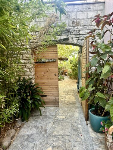 A pathway leading through an archway with wooden doors into a lush garden area, featuring greenery and plants on either side, with a sign indicating a restaurant.