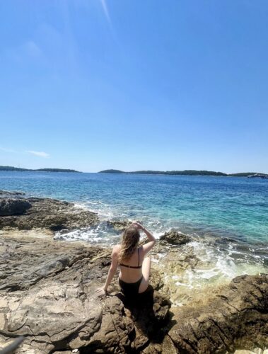 A person sitting on rocky shoreline, enjoying the view of the clear blue sea and distant islands under a sunny sky.