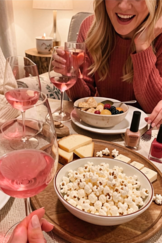 A cozy gathering featuring friends enjoying snacks and drinks, including popcorn, cheese, and a fruit bowl, with pink beverages in glasses.