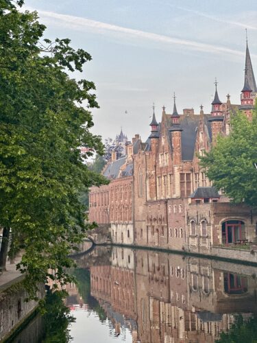 A scenic view of historic buildings along a canal in Bruges, Belgium, with reflection in the water and trees in the foreground.